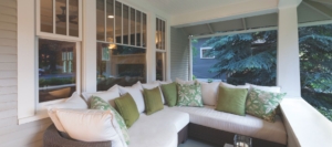 Cozy covered porch with a wicker sectional sofa and green pillows, set against white-framed windows and lush outdoor greenery