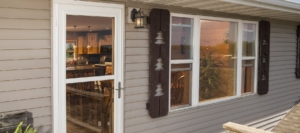 Exterior view of a home showing a glass storm door and windows with tree-shaped cutout shutters, revealing a warm kitchen interior